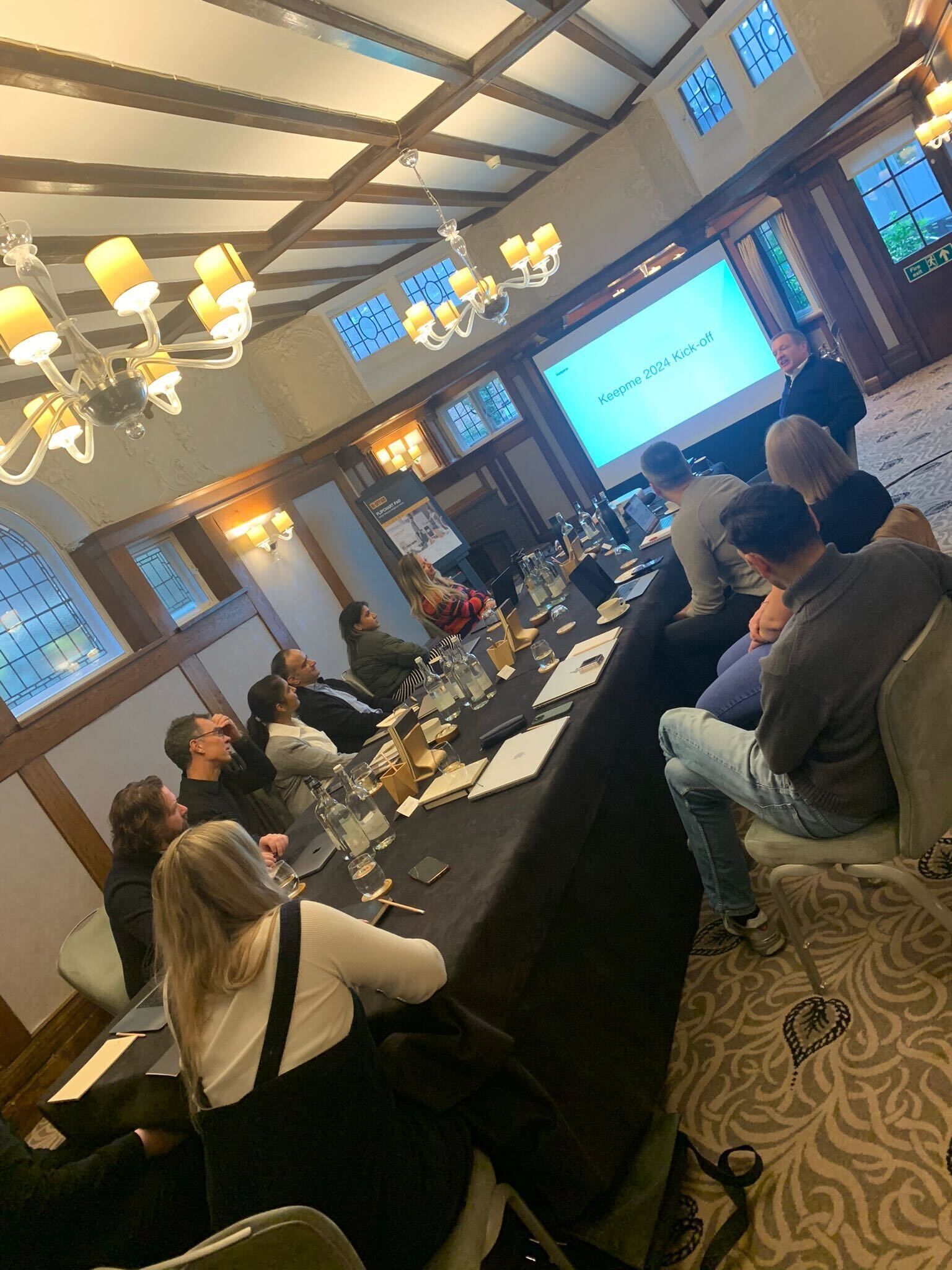 Keepme team members seated at a long table listening to a presentation titled Keepme 2024 Kick-off